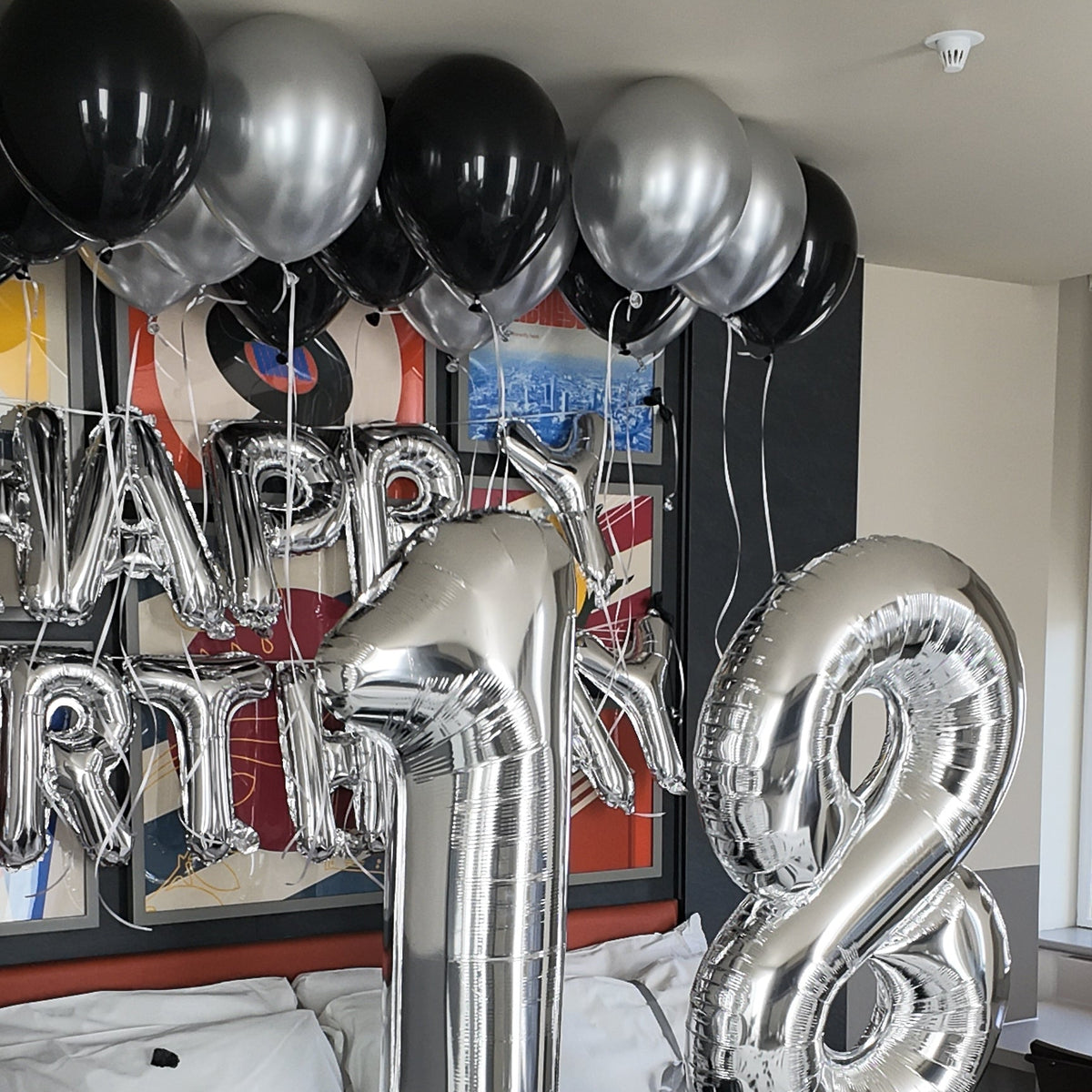 Silver and black foil balloons arranged on a bed, including large number 18 balloons and smaller black balloons, with a 'Happy Birthday' balloon backdrop and confetti scattered on the bedding.