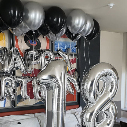 Silver and black foil balloons arranged on a bed, including large number 18 balloons and smaller black balloons, with a 'Happy Birthday' balloon backdrop and confetti scattered on the bedding.